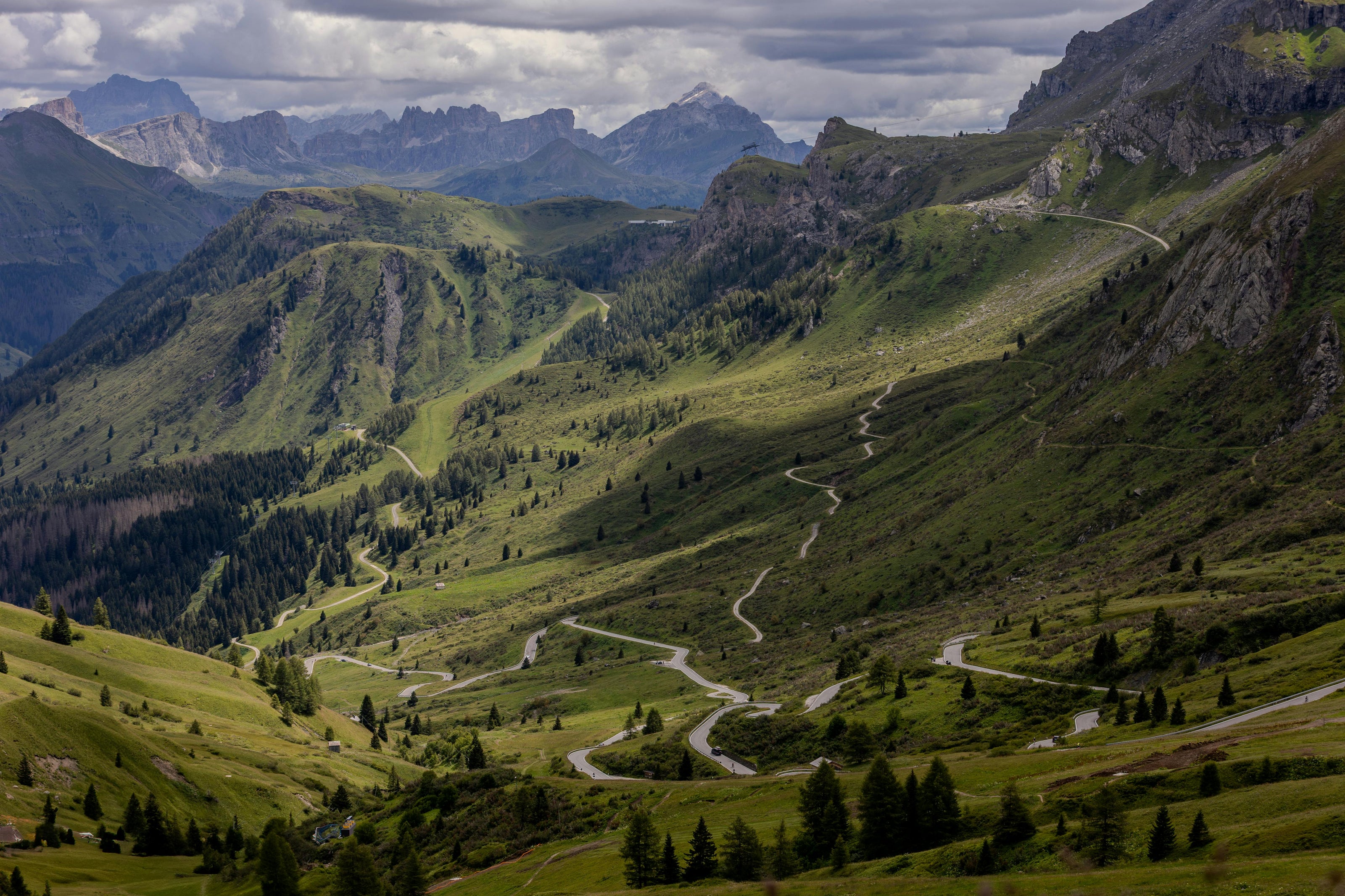 Winding mountain road through a valley with green hills and mountains under a cloudy sky.